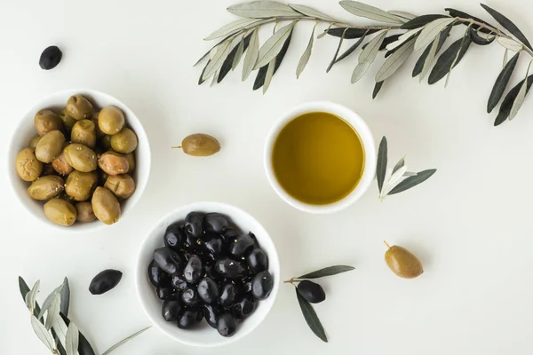 Green and black olives in white bowls next to a bottle with olive oil and leaves on a white background.
