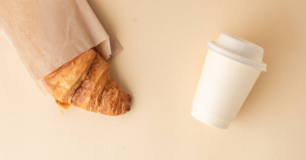 Top view of freshly baked croissant with golden crust next to a disposable paper cup of coffee. French traditional pastry on the beige background. Breakfast in the morning concept