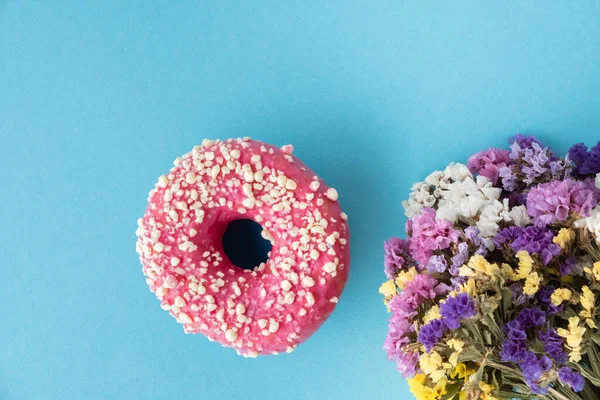 Top view of donut with pink chocolate icing next to bouquet of flowers on the blue pastel background. Mockup with copy space for free text. Flat lay. Donut covered with pink glaze. Spring time concept