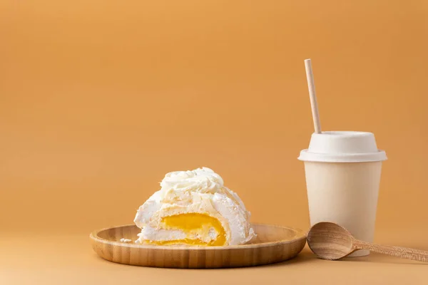 Meringue roll filled with mango puree on the brown background. Meringue roulade with yellow fruit filling in the bamboo wooden plate next to beige paper cup of coffee or tea. Sweet pastry concept