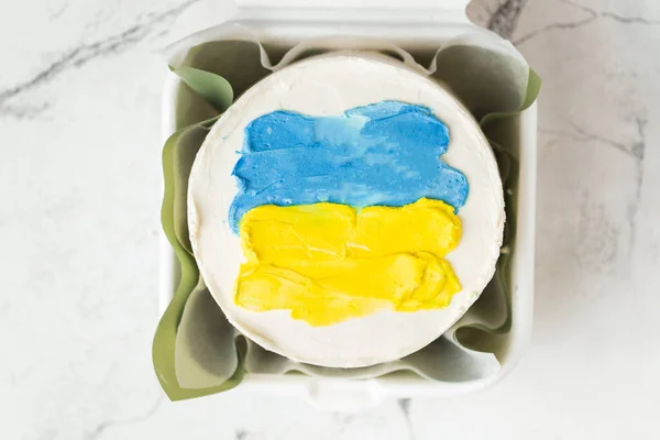 Patriotic cake decorated with yellow and blue Ukrainian flag. Small trendy bento cake in the white gift box. Korean style little cake on the white marble background.
