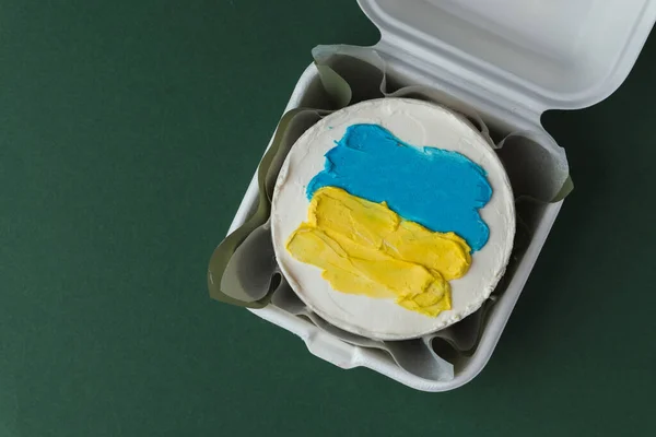 Patriotic cake decorated with yellow and blue Ukrainian flag. Small trendy bento cake in the white gift box. Korean style little cake on the dark green background.