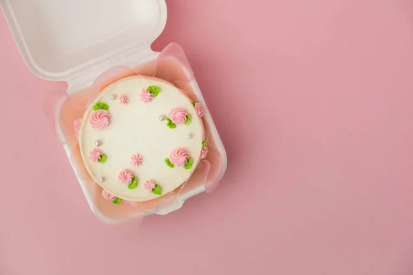Small traditional Korean style bento cake with white cream cheese frosting decorated with pink whipped cream flowers. Little spring cake in the white gift box on the pink pastel background. Top view