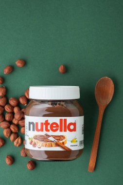 KYIV, UKRAINE - February 21: Nutella chocolate hazelnut spread in a glass jar with white cap next to wooden spoon and raw hazelnuts scattered on the green background. Top view. Copy space.