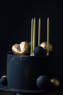 Luxury anniversary cake with black velvet sprayed coating decorated with golden chocolate spheres, heart and candles on top. Vintage cake for beloved one on the black background
