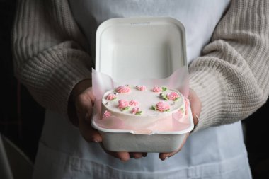Beyaz önlük takan ve krem peynirli pembe çiçeklerle süslenmiş küçük bento pastası tutan bir kadın. Kadın elleri beyaz bir hediye kutusunda doğum günü pastası tutuyor. Modaya uygun Kore tarzı pasta.