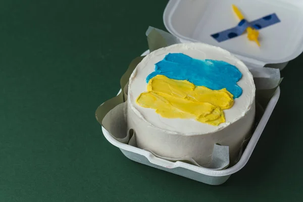 Patriotic cake decorated with yellow and blue Ukrainian flag. Small trendy bento cake in the white gift box. Korean style little cake on the dark green background.