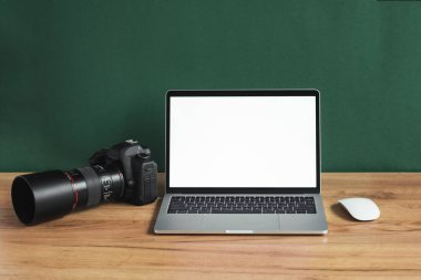 Bilgisayarı, DSLR kamerası ve lensleri olan modern fotoğrafçı işyeri. Koyu yeşil duvara karşı ahşap ofis masasında boş beyaz ekranlı dizüstü bilgisayar. Fotoğraf ya da video yapımcısı reklamı için model