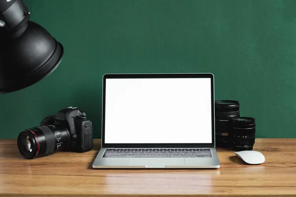 stock image Modern photographer workplace with computer, DSLR camera and lenses. Laptop with the blank white screen on the wooden office table against dark green wall. Mockup for photo or videomaker advertisement