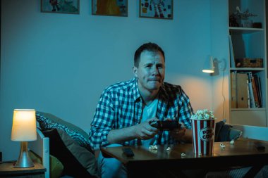 Büyük ekranda video oyunu oynayan ve evde patlamış mısır yiyen konsantre beyaz bir adam. Geceleri mavi ekran ışığıyla dolu bir odada elinde oyun konsolunun kablosuz kumandanı olan bir adam..