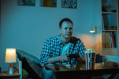 Büyük ekranda video oyunu oynayan ve evde patlamış mısır yiyen konsantre beyaz bir adam. Geceleri mavi ekran ışığıyla dolu bir odada elinde oyun konsolunun kablosuz kumandanı olan bir adam..