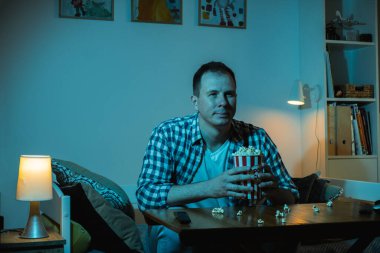 Adam TV izliyor ve gece evde patlamış mısır yiyor. Bir kanepede oturan bir adamın portresi. Kocaman bir televizyonun mavi ekran ışığıyla dolu bir odada.