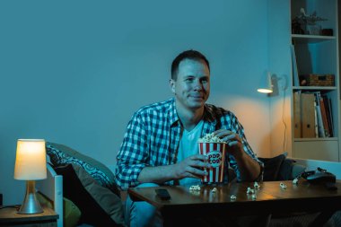 Adam TV izliyor ve gece evde patlamış mısır yiyor. Bir kanepede oturan bir adamın portresi. Kocaman bir televizyonun mavi ekran ışığıyla dolu bir odada.