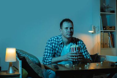 Adam TV izliyor ve gece evde patlamış mısır yiyor. Bir kanepede oturan bir adamın portresi. Kocaman bir televizyonun mavi ekran ışığıyla dolu bir odada.