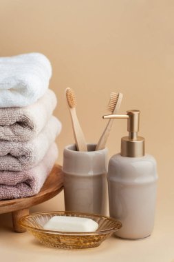 Sabun ve diş fırçalarının yanında bir yığın temiz yumuşak havluyla klasik altın banyo malzemeleri. Beyaz, pembe ve bej havlular. Bej arka planda banyo aksesuarları var..