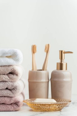 Sabun ve diş fırçalarının yanında bir yığın temiz yumuşak havluyla klasik altın banyo malzemeleri. Beyaz arka planda banyo aksesuarları olan beyaz, pembe ve bej havlular..