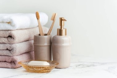 Sabun ve diş fırçalarının yanında bir yığın temiz yumuşak havluyla klasik altın banyo malzemeleri. Beyaz arka planda banyo aksesuarları olan beyaz, pembe ve bej havlular..