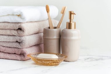 Sabun ve diş fırçalarının yanında bir yığın temiz yumuşak havluyla klasik altın banyo malzemeleri. Beyaz arka planda banyo aksesuarları olan beyaz, pembe ve bej havlular..