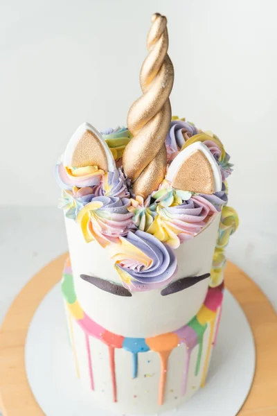 Big bunk cake with white chocolate frosting decorated with colorful drips, mastic eyebrows and golden ears with horn on top. Birthday party unicorn cake for a little girl on the white background.