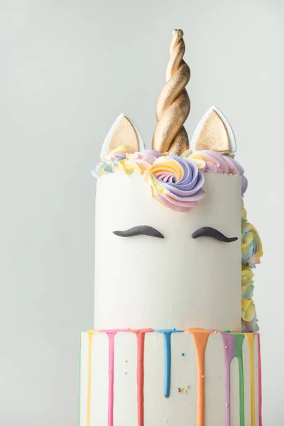 Big bunk cake with white chocolate frosting decorated with colorful drips, mastic eyebrows and golden ears with horn on top. Birthday party unicorn cake for a little girl on the white background.