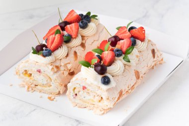 Beyaz arka plandaki beyaz hediye kutusunda badem gevreği, yaban mersini ve çilekle süslenmiş ev yapımı krema rulosu. Krem şantili kremalı krema..