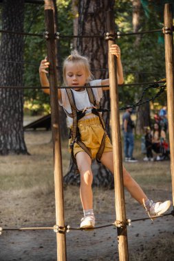 Şirin küçük kız tırmanır ve ahşap bir patikada yürür ve uzun yeşil kozalaklı ağaçların yüksek ipleri ile ip parkında durur. Macera halat parkında eğlenirken güvenlik önlemleri