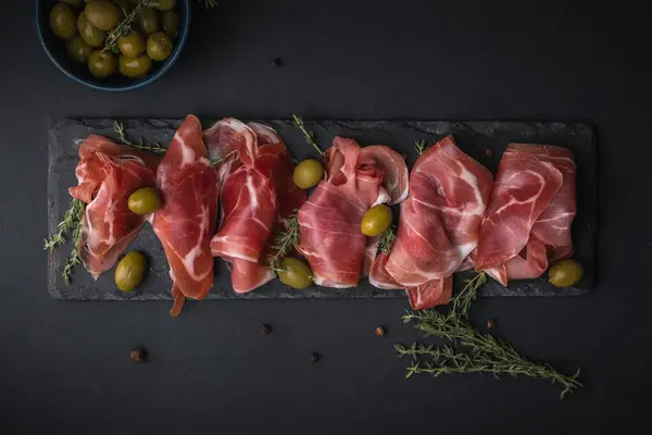 Kurutulmuş Serrano jambonu ya da İspanyol jamonu iberico 'dan aperatifler. İtalyan jambonlu, yeşil zeytinli ve siyah taşlı. Siyah arkaplanda et dilimleri