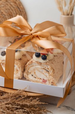 Bej arka planda şeffaf hediye kutusunda badem gevreği, yabanmersini ve çilekle süslenmiş ev yapımı krema rulosu. Krem şantili kremalı krema..