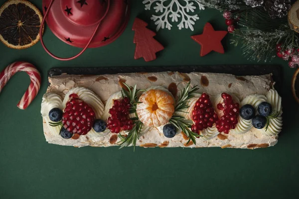 Meringue roll sprinkled with powdered sugar decorated with blueberries, pomegranate and almond flakes on the dark green Christmas festive background. Flat lay