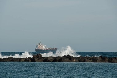 Dalgalar okyanusta dalgalanan sulara çarpıyor ve arka planda bir gemi var. Dalgalar fren yapar ve kayaya çarpar. Uzakta büyük bir gemi ücretsiz metin için bir kopya alanı ile