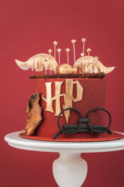 KYIV, UKRAINE - March 03: Harry Potter cake on the burgundy red background. Birthday magic cake with red velvet chocolate coating decorated with mastic glasses, Elder Wand and Golden Snitch clipart