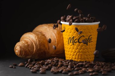 KYIV, UKRAINE - Ocak 06: Popüler bir fast food restoranında taze kruvasanla sarı McDonald 's kağıt bardağına yakın çekim yapın. Siyah arka planda uçan kahve çekirdekleri olan McCafe bardağı.