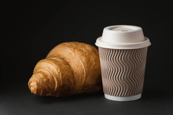 Paper cup of hot coffee with paper lid next to freshly baked croissant on the black background. Mock up with copy space for a free text