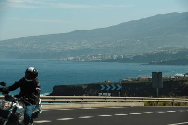 Arka planda büyük dağlar olan deniz kenarındaki yolun güzel manzarası. Motosikletli, deniz kenarındaki otoyolda. Motosiklet konseptiyle yolculuk