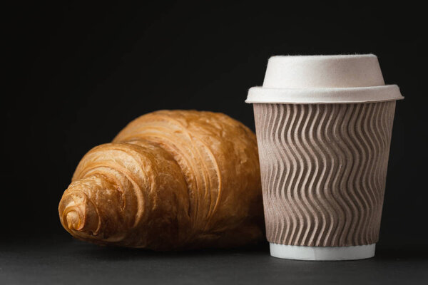 Paper cup of hot coffee with paper lid next to freshly baked croissant on the black background. Mock up with copy space for a free text