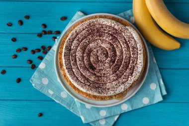 Top view of banoffe pie filled with banana and dulce de leche and sprinkled with grated chocolate. Banana cake on white plate with glass of coffee on turquoise summer background. Flat lay clipart