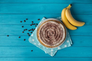 Top view of banoffe pie filled with banana and dulce de leche and sprinkled with grated chocolate. Banana cake on white plate with glass of coffee on turquoise summer background. Flat lay clipart