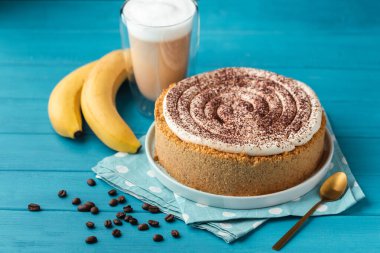 Banoffe pie filled with banana and dulce de leche and sprinkled with grated chocolate. Banana cake on white plate with glass of coffee on turquoise summer background clipart