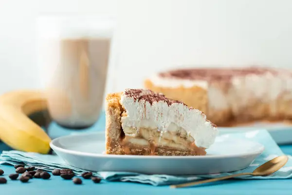 stock image Slice of banoffe pie filled with banana and dulce de leche and sprinkled with grated chocolate. Banana cake on white plate with glass of coffee on turquoise summer background