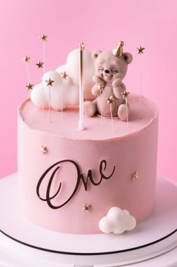 Oyuncak ayı ve beyaz bulutlarla süslenmiş pembe çikolata kaplı küçük bir bebek için doğum günü pastası. Pembe arka planda küçük bir kız için doğum günü pastası