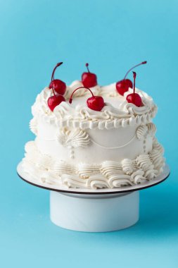 Delicate lambeth style birthday cake with white cream cheese frosting decorated with cocktail cherries on the blue background clipart