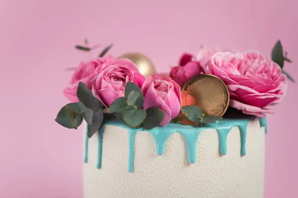 Anniversary cake with white chocolate velvet coating decorated with fresh pink roses and turquoise chocolate drips. Birthday cake for a girl on a pink background