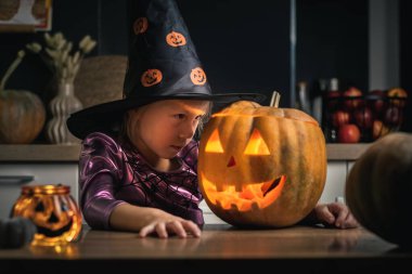 Ürkütücü, parıldayan Cadılar Bayramı balkabağının yanında, ürkütücü, şenlikli cadı kıyafetli tatlı kız. Küçük kız mutfakta masada otururken Jack-o-fener yapıyor. Mutlu Cadılar Bayramı