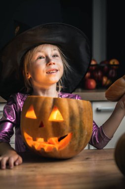 Ürkütücü, parıldayan Cadılar Bayramı balkabağının yanında, ürkütücü, şenlikli cadı kıyafetli tatlı kız. Küçük kız mutfakta masada otururken Jack-o-fener yapıyor. Mutlu Cadılar Bayramı