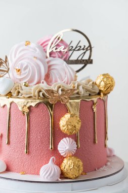 Güzel lüks doğum günü pastası pembe çikolata kremalı altın renkli çikolata damlaları ve beyaz arka planda altın şekerlerle süslenmiş.