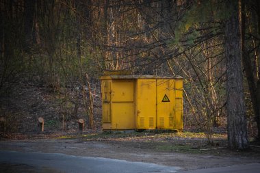 Ormanda yüksek voltajlı transformatör kabini. Kırsal bölgedeki sarı trafo evinin kapısında yüksek voltaj işareti var.