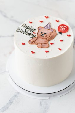 Oyuncak ayının balon çizimiyle süslenmiş şirin bir doğum günü pastası. Beyaz mermer arka planda mutlu yıllar mesajı olan beyaz pasta.