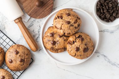 Beyaz mermer arka planda fındıklı ve çikolata damlacıklı taze çikolatalı Amerikan kurabiyeleri. Modaya uygun çatal bıçak takımları ve pasta şefi aksesuarları olan düz bir kurabiye. Ev yapımı kurabiye tarifi
