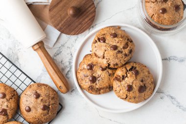 Beyaz mermer arka planda fındıklı ve çikolata damlacıklı taze çikolatalı Amerikan kurabiyeleri. Modaya uygun çatal bıçak takımları ve pasta şefi aksesuarları olan düz bir kurabiye. Ev yapımı kurabiye tarifi