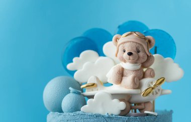 Big two tier birthday cake with blue chocolate frosting decorated with teddy bear flying among the clouds on the plane figurine. Beautiful birthday cake for a little one year old boy. Blue background